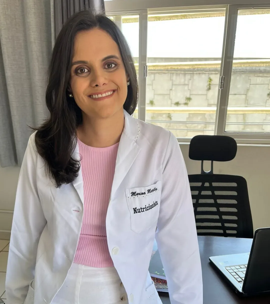 Marina Meirelles, nutricionista, sorrindo, de jaleco branco e blusa rosa no consultório